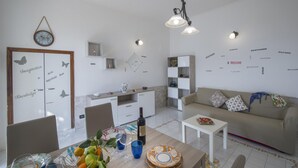 A modern living room with a sofa, a dining table with fruit and wine, and a kitchen area with a fridge and cabinets.