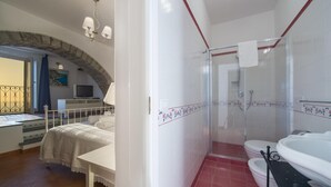 Une chambre avec un lit, une télévision et une arche en pierre. La salle de bain est équipée d’une douche, d’une baignoire et de toilettes.