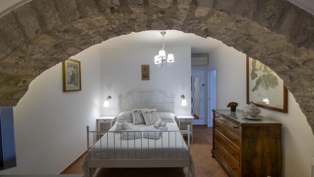 Une chambre avec une arche en pierre, un lit recouvert de draps blancs, des lampes de chevet, une commode et un tableau accroché au mur.
