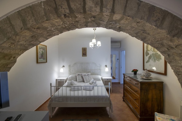 A bedroom with a stone arch, a bed with white linens, bedside lamps, a dresser, and a painting on the wall.