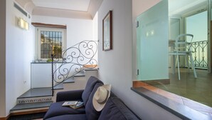 Un salon avec un canapé foncé, un chapeau posé sur l’accoudoir, un escalier orné d’une rampe décorative et une vue sur un balcon avec une chaise.
