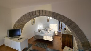 Une chambre à coucher avec une arche en pierre, un lit, un téléviseur posé sur un meuble blanc et une table de chevet avec une lampe.