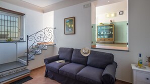 Un salon avec un canapé, un tableau encadré au mur, un escalier avec une rampe en métal et un coin cuisine visible à travers une fenêtre.