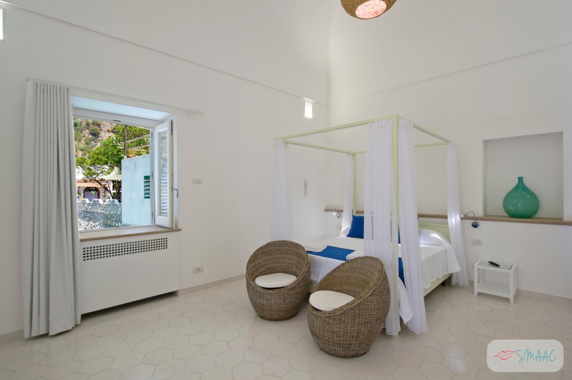 Une chambre à coucher avec un lit à baldaquin, deux fauteuils en osier, une petite table de chevet et une vue sur l’extérieur par une fenêtre.