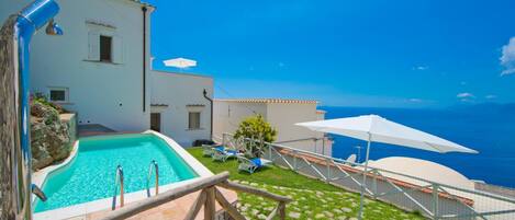 A pool area with a clear view of the sea, a white building with a tiled roof, and a wooden deck.