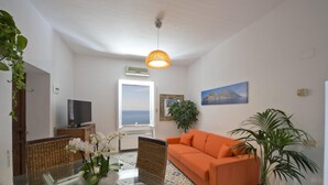 Een woonkamer met een bank, een televisie, een raam met uitzicht op zee en een potplant.