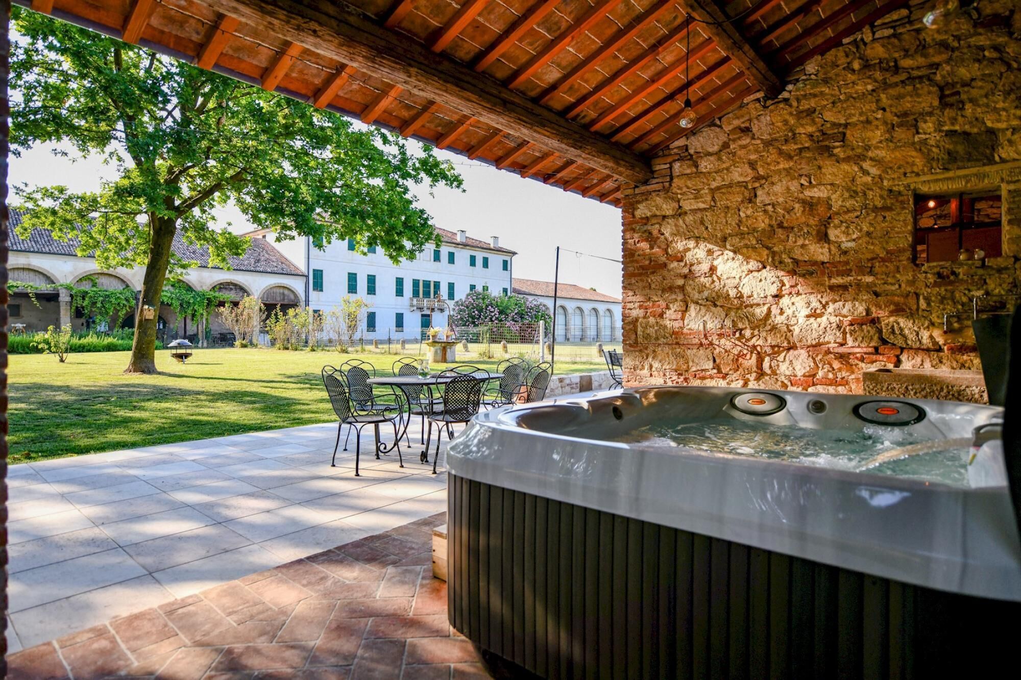 Un bain à remous sous une pergola en bois, avec un mur en pierre en arrière-plan, des sièges d’extérieur et une vue sur un bâtiment doté de fenêtres en arc de cercle.