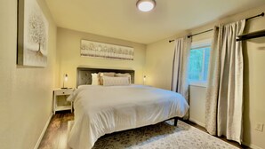 A bedroom with a large bed, a nightstand with a lamp, a wall-mounted artwork, and a window with curtains. - Rustic, pet-friendly Olympia cottage on wooded acres with patio, fire pit, and modern comforts (Olympia)