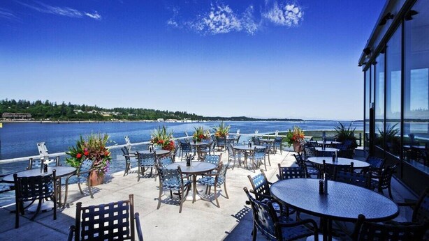 An outdoor patio with round tables and chairs, overlooking a body of water and a distant shoreline. - Rustic, pet-friendly Olympia cottage on wooded acres with patio, fire pit, and modern comforts (Olympia)