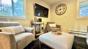 A living room with two armchairs, a coffee table, a wall clock, and a TV. - Rustic, pet-friendly Olympia cottage on wooded acres with patio, fire pit, and modern comforts (Olympia)