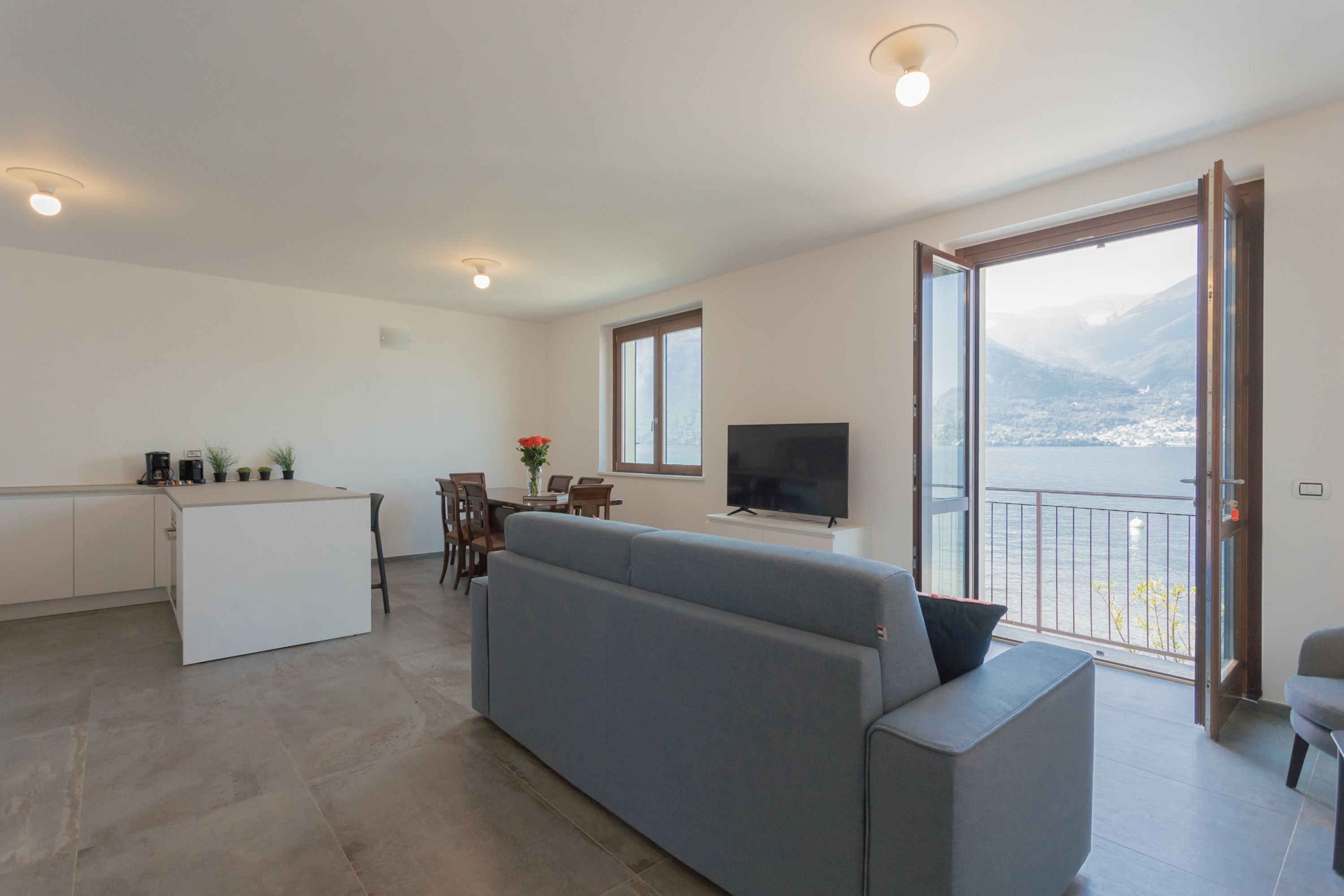 A modern living room with a grey sofa, a TV, and a balcony with a view of a city and mountains.