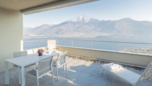 Ein Balkon mit Tisch, Stühlen und einer Liege, mit Blick auf Berge und ein Gewässer.