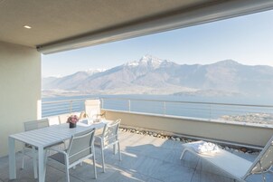 Un balcón con mesa y sillas, una silla de descanso y vistas a montañas y una masa de agua.