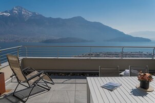 Un balcón con vista a un lago y montañas, con una mesa, una revista y un florero.