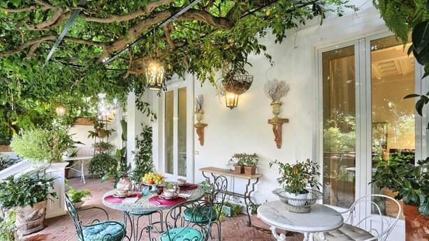 Un patio al aire libre con mesas y sillas, rodeado de plantas en macetas y plantas colgantes.