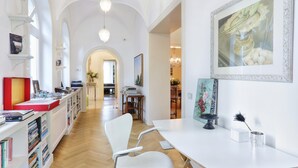 Una habitación amplia con techo alto, un escritorio blanco con silla, estanterías y un cuadro grande con marco.