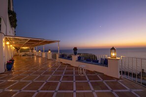 Eine Terrasse mit gefliestem Boden, Sitzgelegenheiten im Freien und Blick auf das Meer bei Sonnenuntergang.