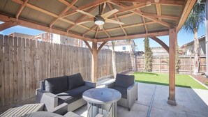 Een overdekt terras met een plafondventilator, tuinmeubilair en een houten omheining op de achtergrond.