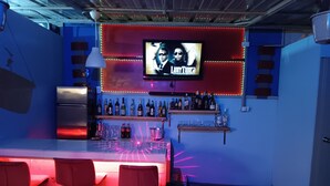 Een bar met een televisiescherm waarop een programma draait, planken vol flessen en rode barstoelen.