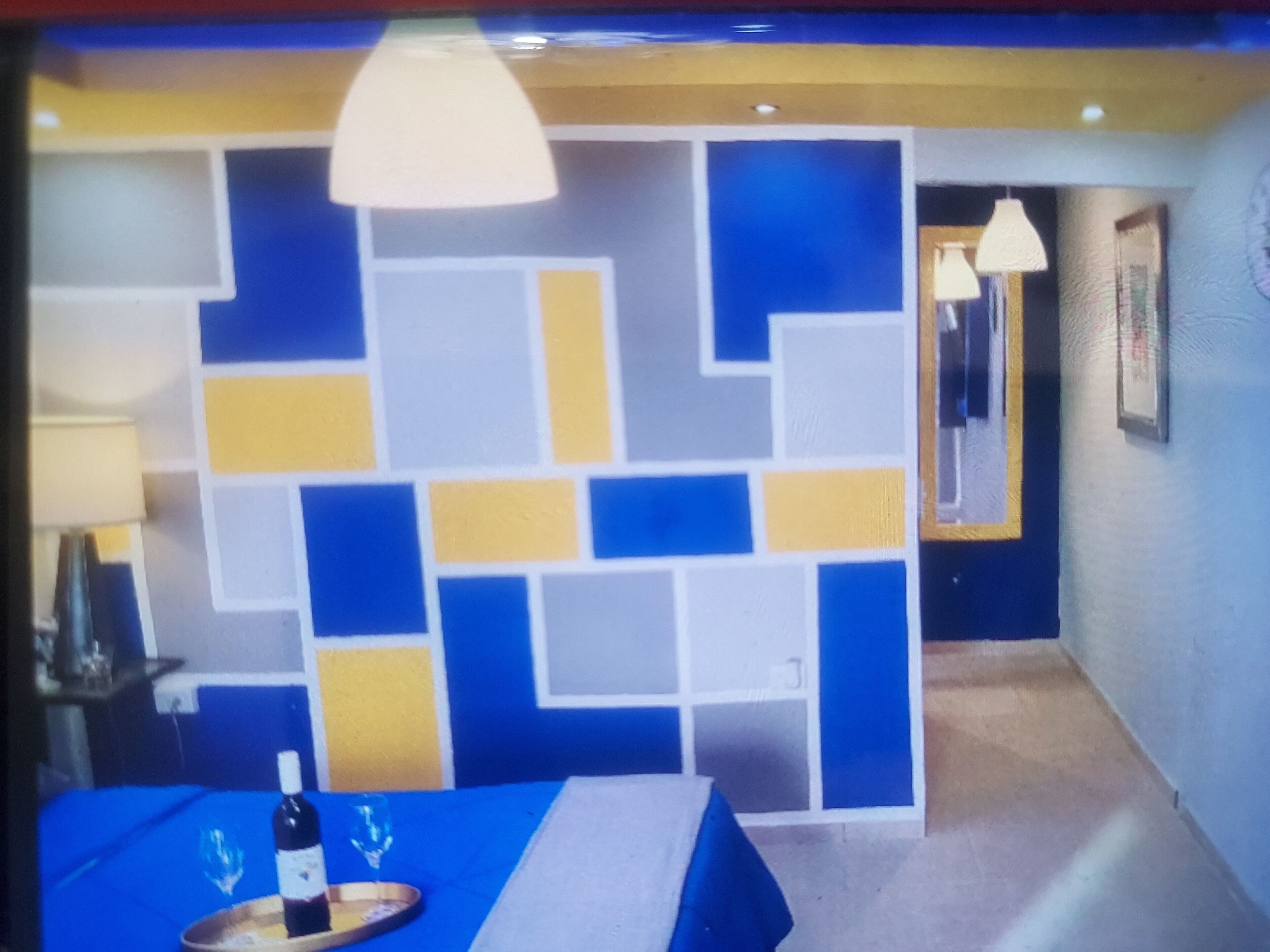 Een hotelkamer met een wand met geometrisch patroon, een bed met een blauwe spreideken, een nachtkastje met een fles wijn en een glas, en een lamp.