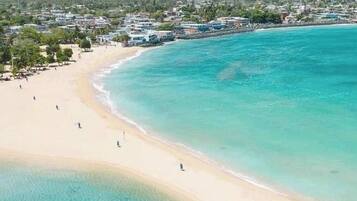 An aerial view of a beach with clear turquoise waters, a sandy shore, and buildings in the background.