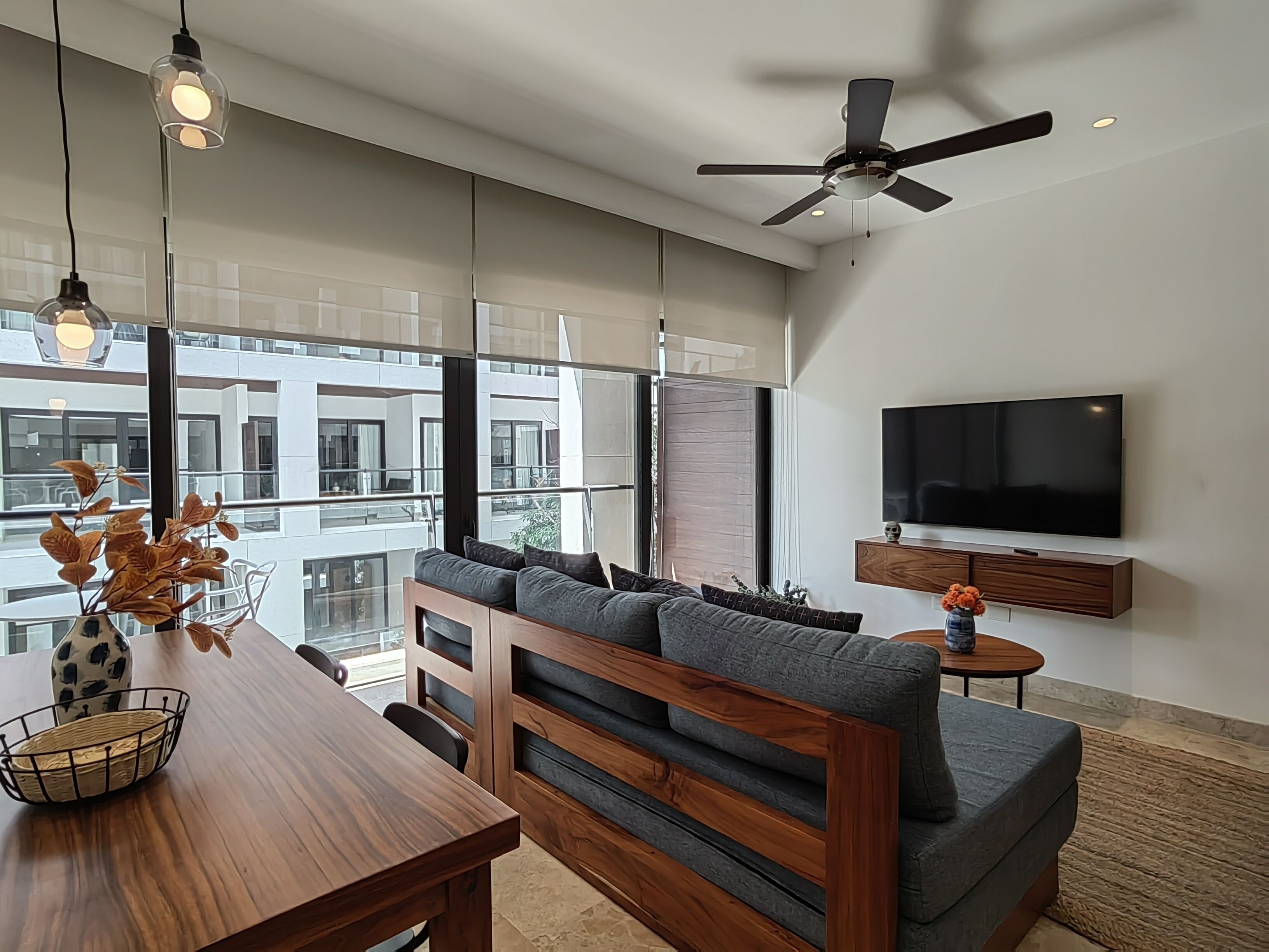 A modern living room with a wooden dining table, grey sofa, and a ceiling fan. A flat-screen TV is mounted on the wall.