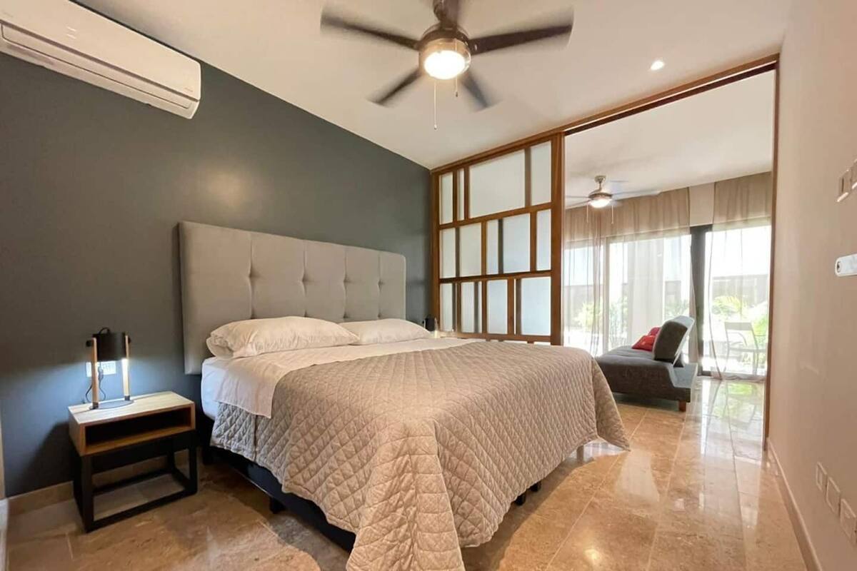 A modern bedroom with a large bed, a nightstand, a ceiling fan, and a view of the outdoors through large windows.