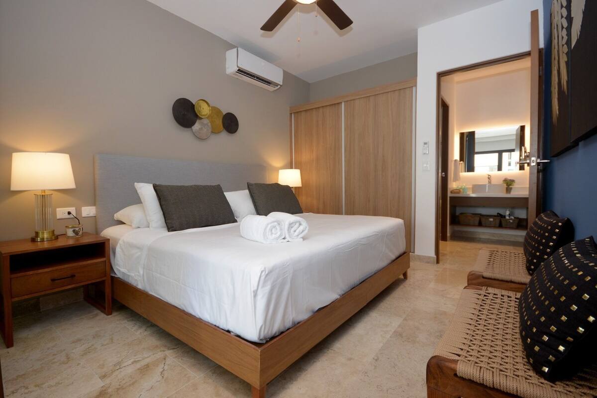 A hotel room with a bed, bedside lamps, a nightstand, a wooden wardrobe, a bathroom with a sink and mirror, and a ceiling fan.