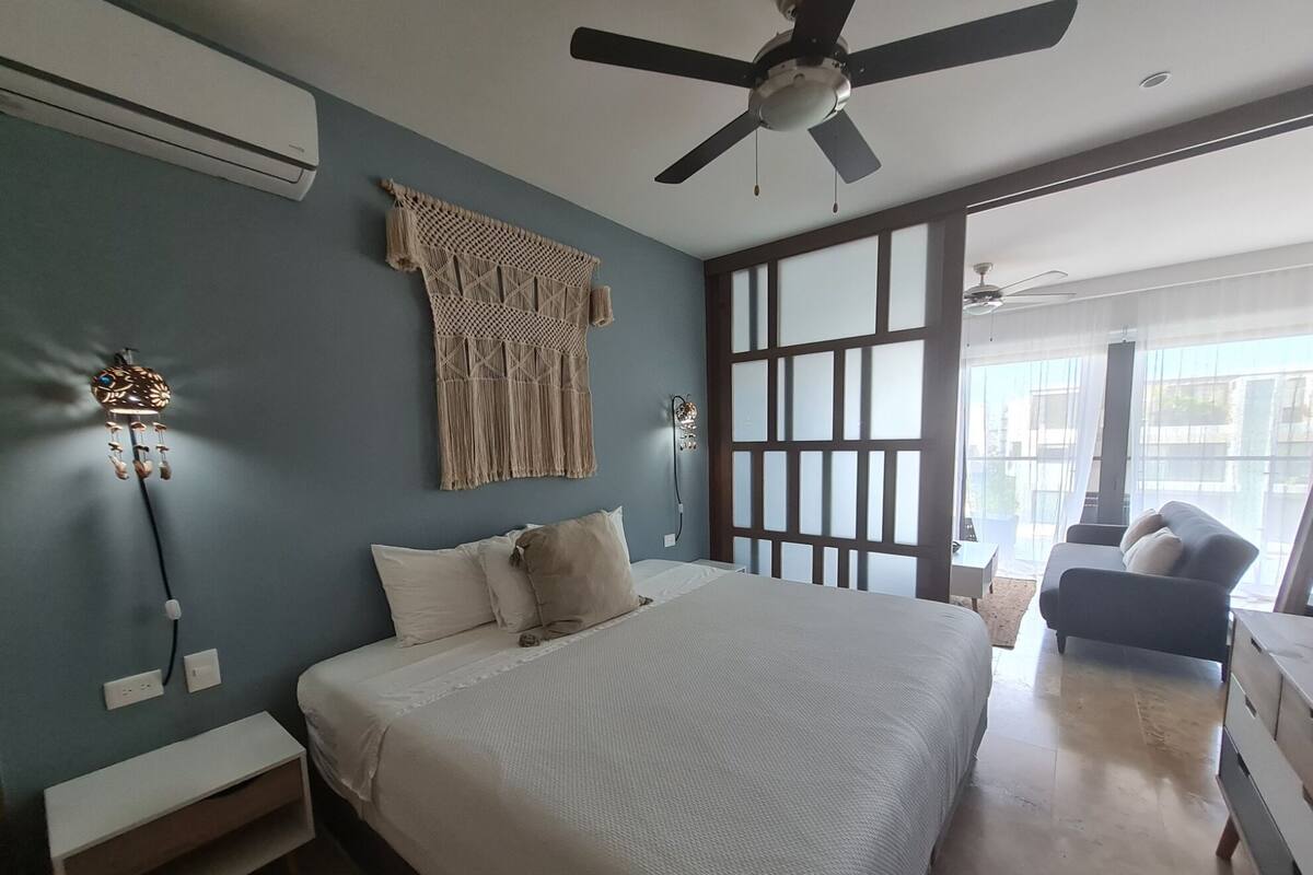 A bedroom with a bed, a ceiling fan, a macrame wall hanging, and a view through a sliding door.