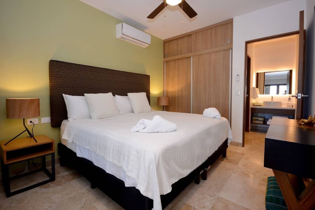 A neatly made bed with white linens and pillows, a bedside table with a lamp, a wooden headboard, and a wardrobe with sliding doors.