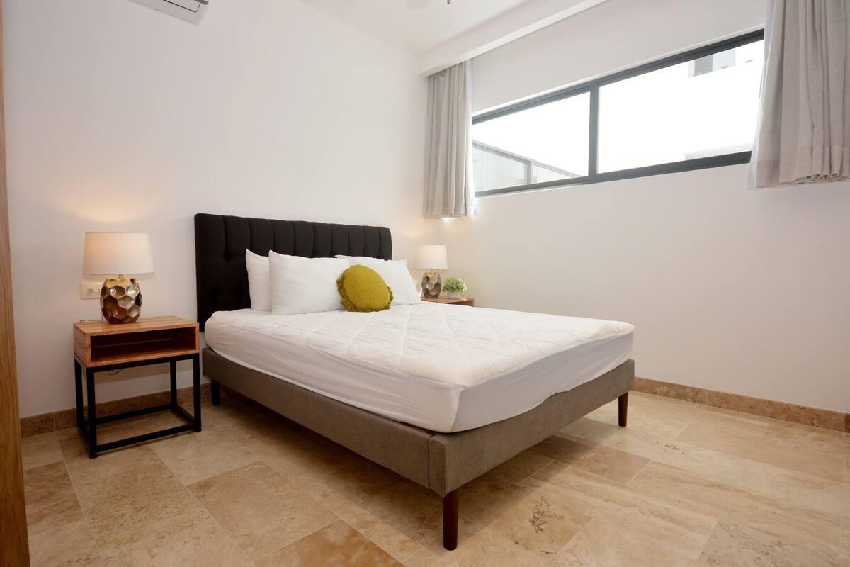 A neatly made bed with a grey headboard and white bedding, two bedside tables with lamps, and a large window with curtains.