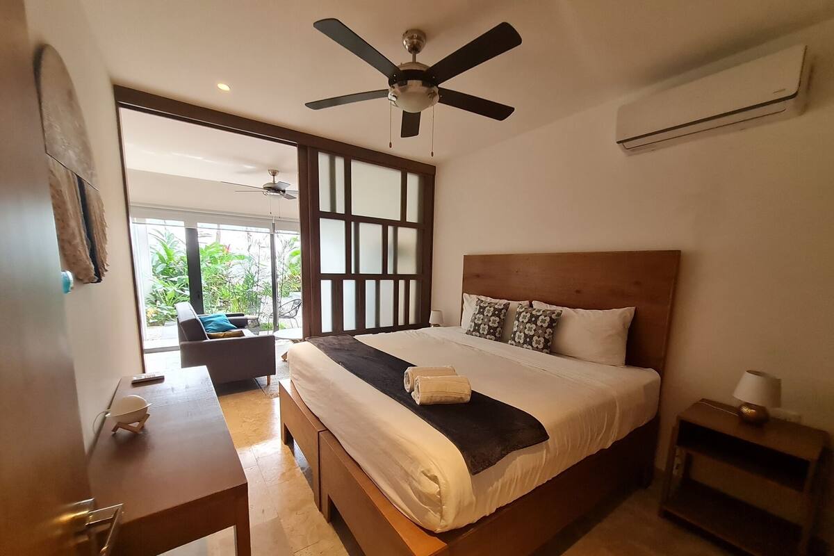 A bedroom with a bed, a desk, a chair, a ceiling fan, and a view of the outdoors.