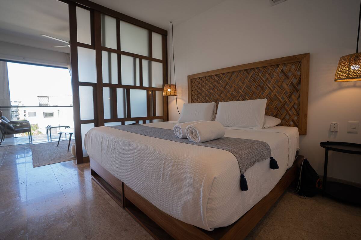A neatly made bed with white linens and a grey throw blanket, a wooden headboard with a woven pattern, a small table with a lamp, and a view of a balcony with outdoor furniture.