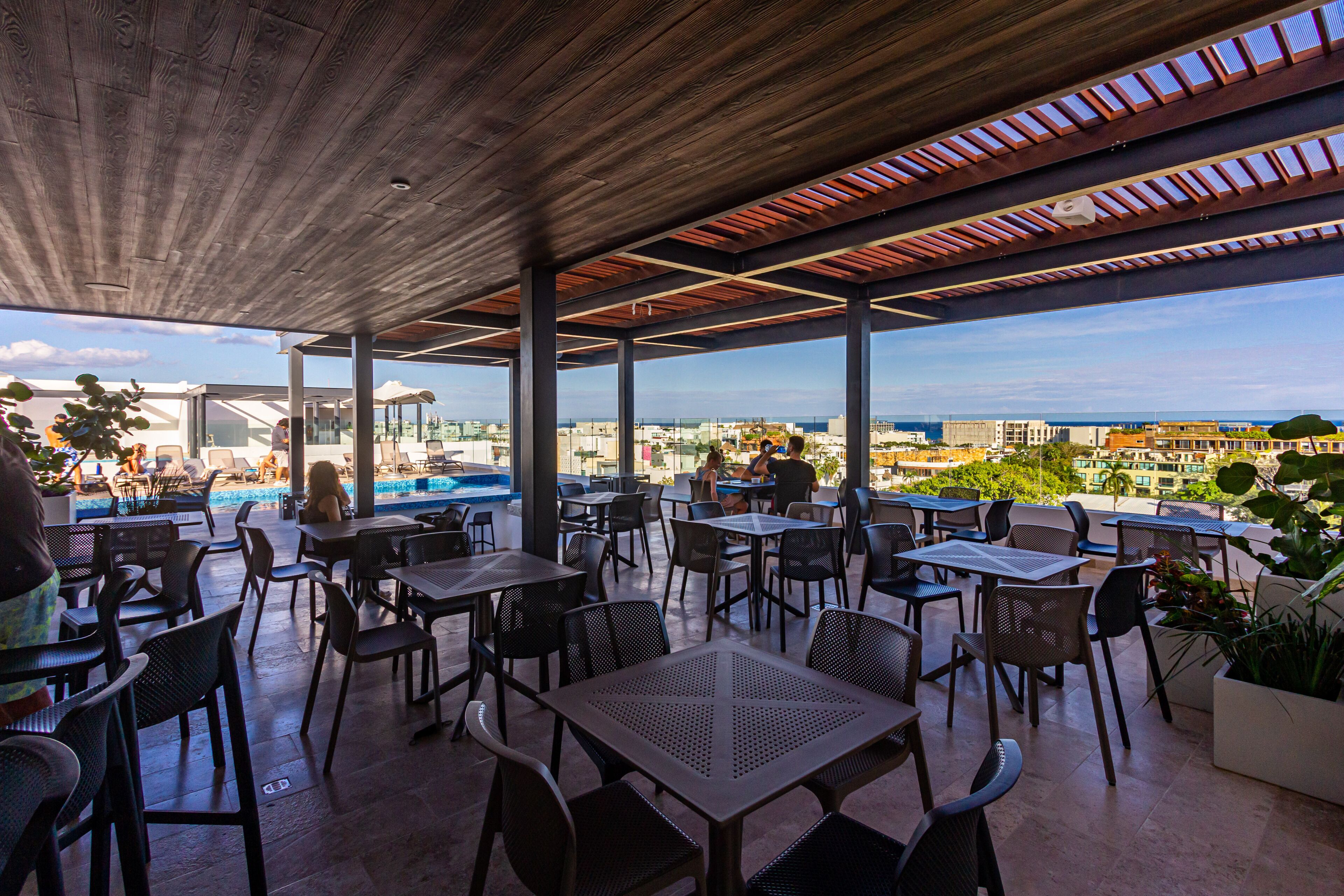 Un patio exterior con mesas y sillas, con vistas a una piscina y al horizonte urbano.