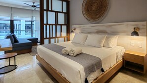 A modern hotel room with a large bed, a ceiling fan, a view of the city, and a small table with a chair. - Ipana Apartment Deluxe (Playa del Carmen)