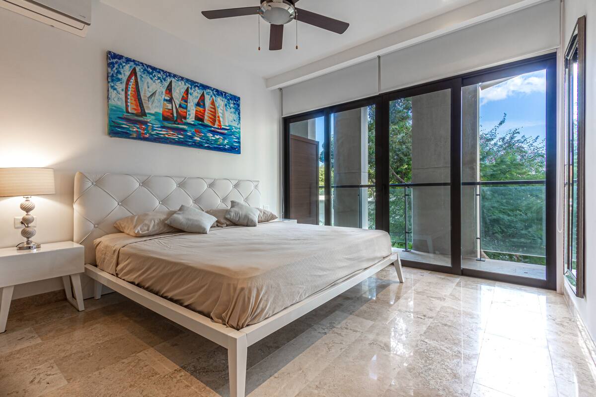 A bedroom with a large bed, a ceiling fan, a painting of sailboats, and a balcony with large windows.