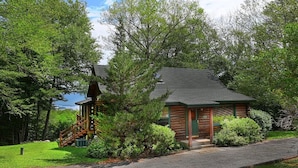 A log cabin with a staircase leading to a deck, surrounded by lush greenery and a view of the water.
