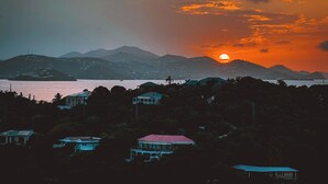 Rannikkomaisema auringonlaskun aikaan, taustalla vuoria ja useita punaisilla kattoilla varustettuja rakennuksia.