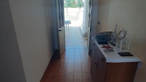 Un couloir étroit, dont le sol est recouvert de carreaux, mène à une porte ouverte. On aperçoit près de la porte une petite table recouverte d’une nappe bleue et une chaise.