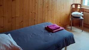 Una habitación con paredes de madera, una cama con colchón azul y manta rosa, una silla con cojín blanco y una ventana con vistas a la vegetación.