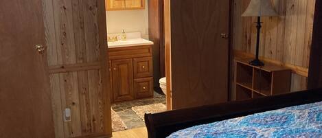 A bedroom with a wooden floor, a bed with a patterned bedspread, a wooden wardrobe, a bathroom with a mirror and sink, and a nightstand with a lamp.