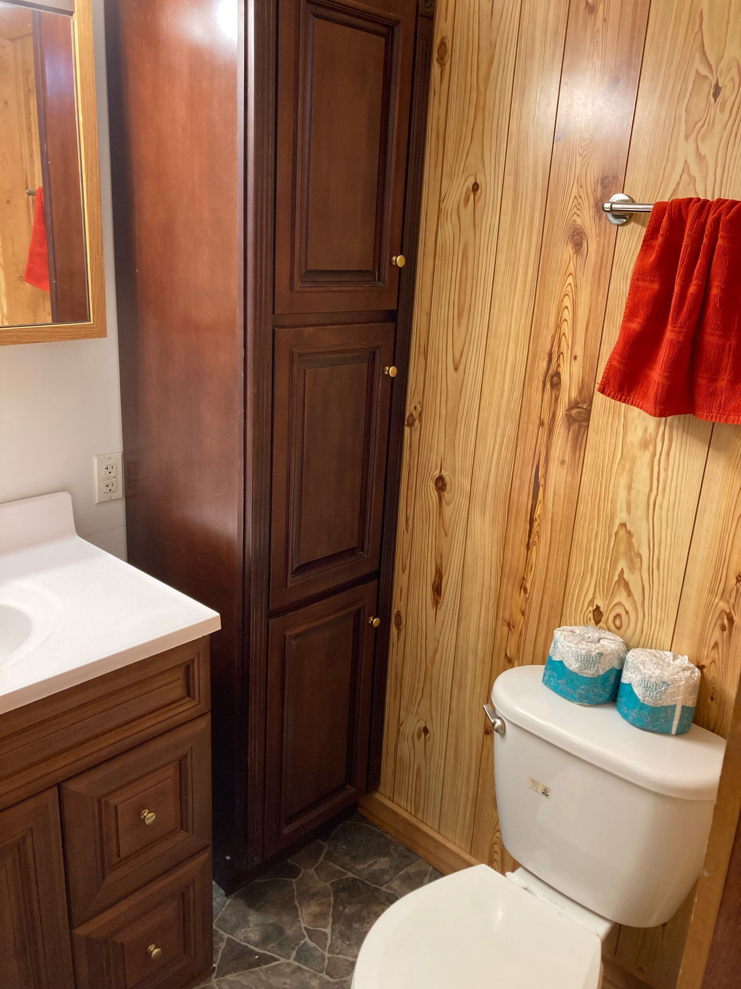 Un baño con tocador de madera, un lavabo blanco, un inodoro con papel higiénico y una toalla roja colgada en la pared.