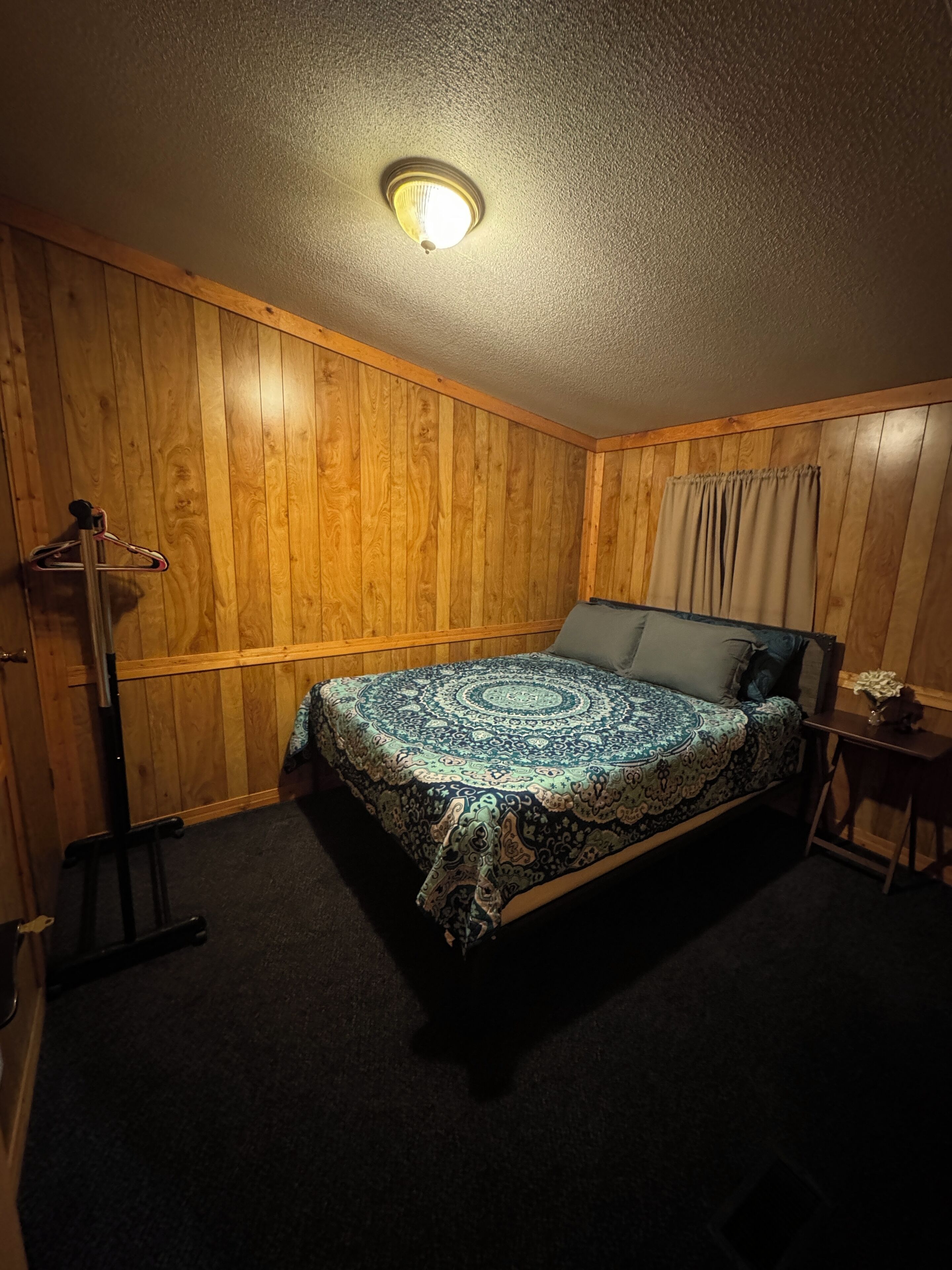 A bedroom with a wooden paneled wall, a bed with a patterned bedspread, a nightstand, a lamp, and a ceiling light.