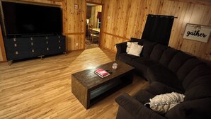 Una sala de estar acogedora con paredes y suelos de madera, un sofá oscuro, una mesa de centro con libros y un vaso, y un televisor.