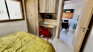 Een slaapkamer met een bed, een bureau en een stoel. Er is een kledingkast, een raam en een deur die leidt naar een andere kamer.
