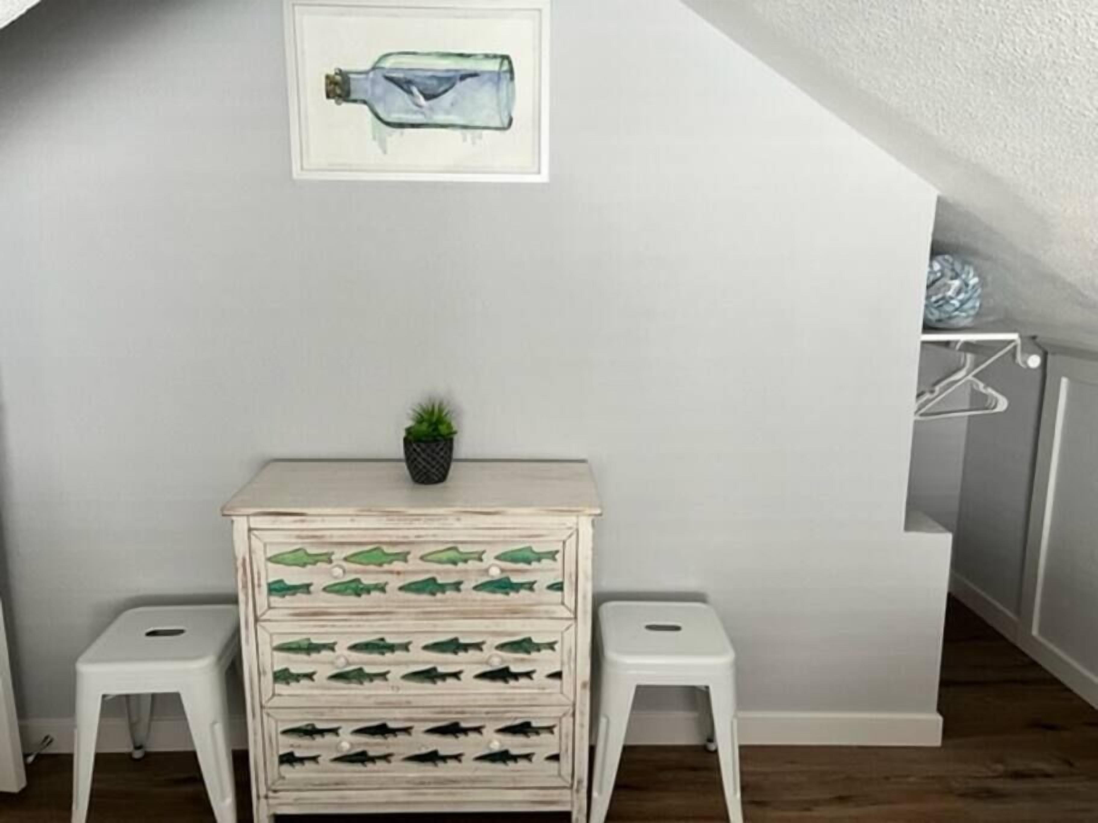 A room with a white wall, a framed picture of a bottle, a small potted plant, and a dresser with drawers decorated with fish.