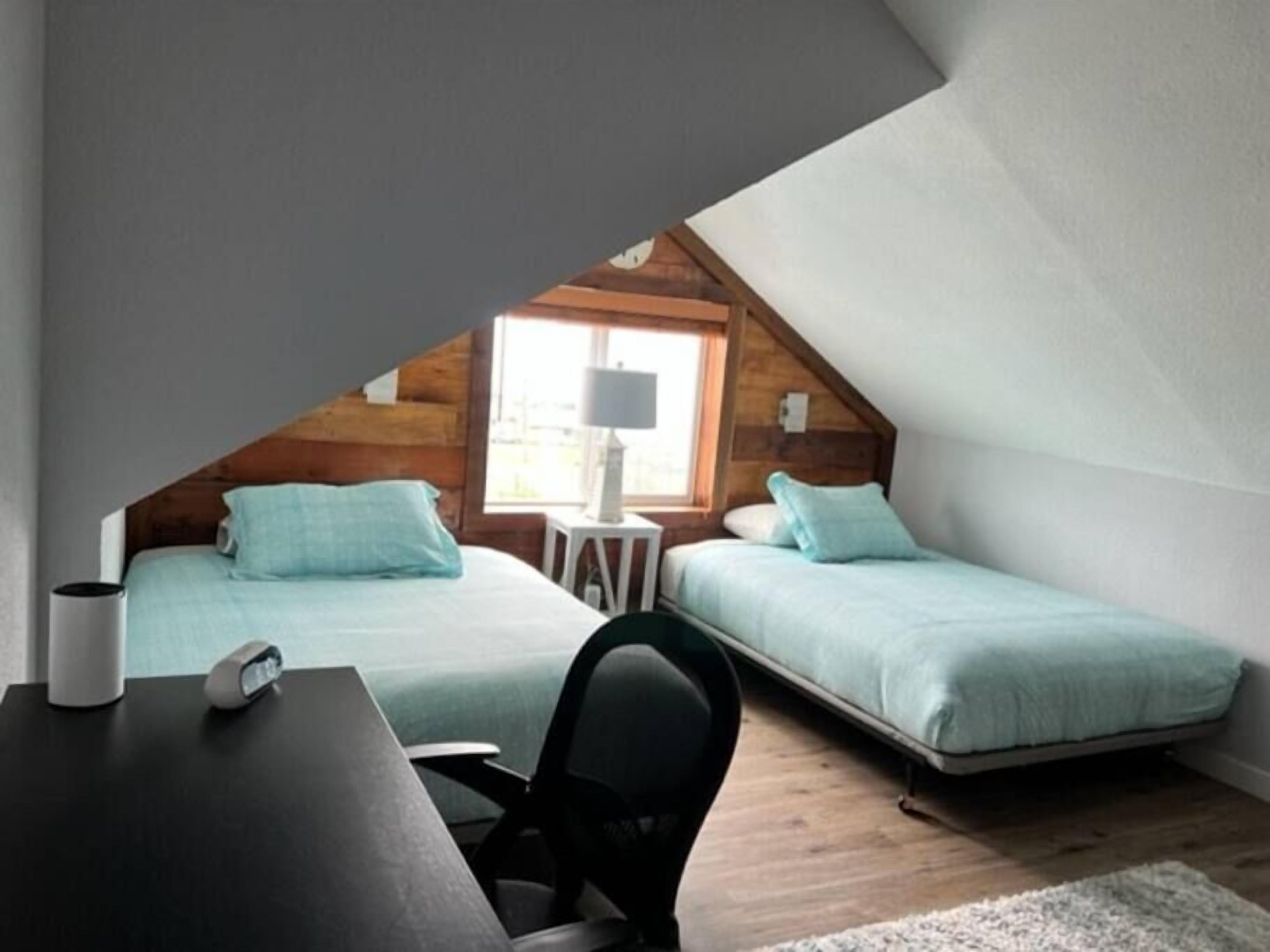 Une petite chambre mansardée, chaleureuse et accueillante, avec deux lits, un bureau et une lampe.