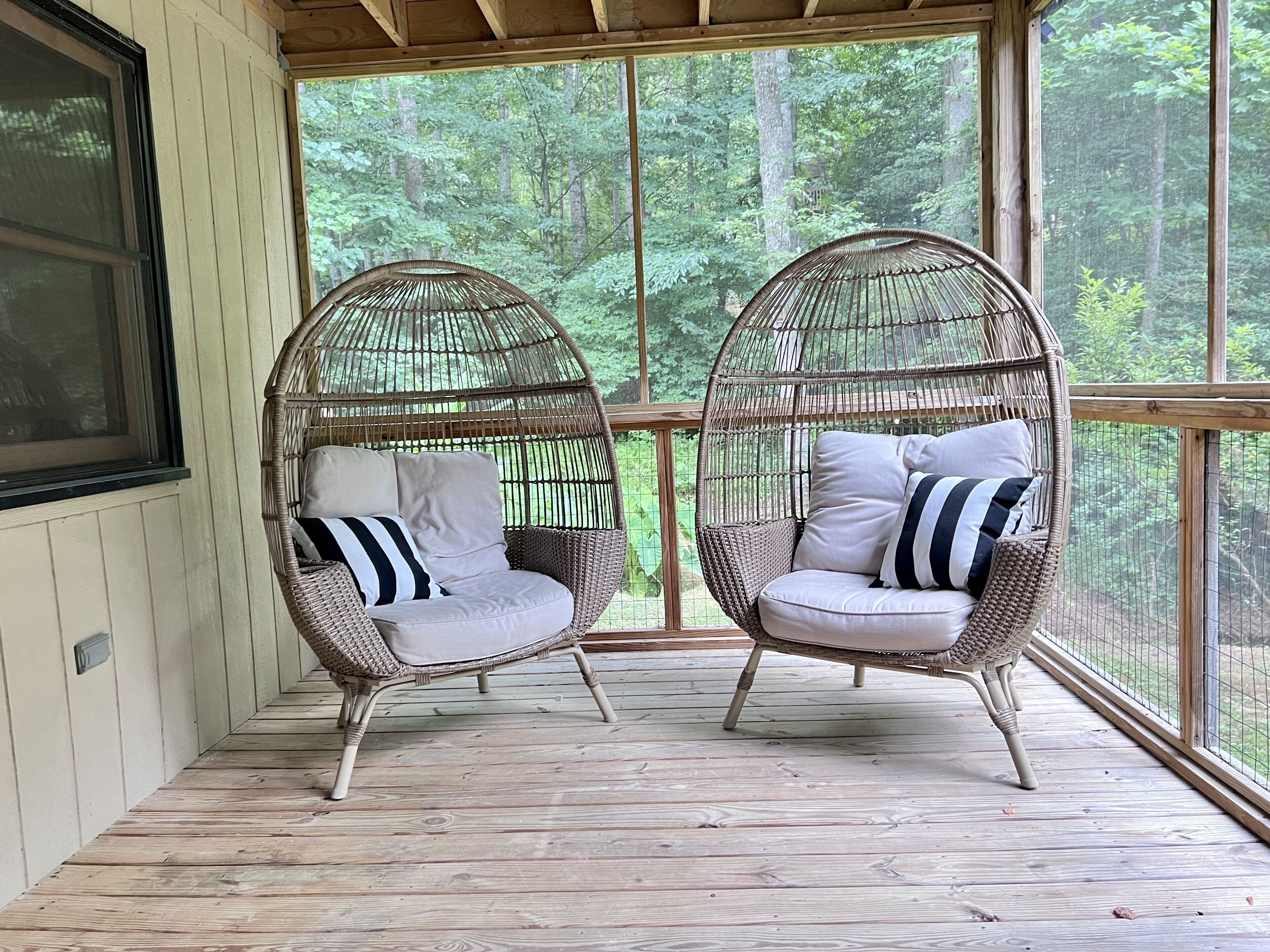 Dos sillas de mimbre con cojines sobre una terraza de madera con amplias ventanas que dan a un bosque.
