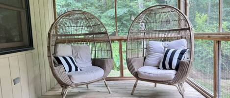 Dos sillas de mimbre con cojines sobre una terraza de madera con amplias ventanas que dan a un bosque.