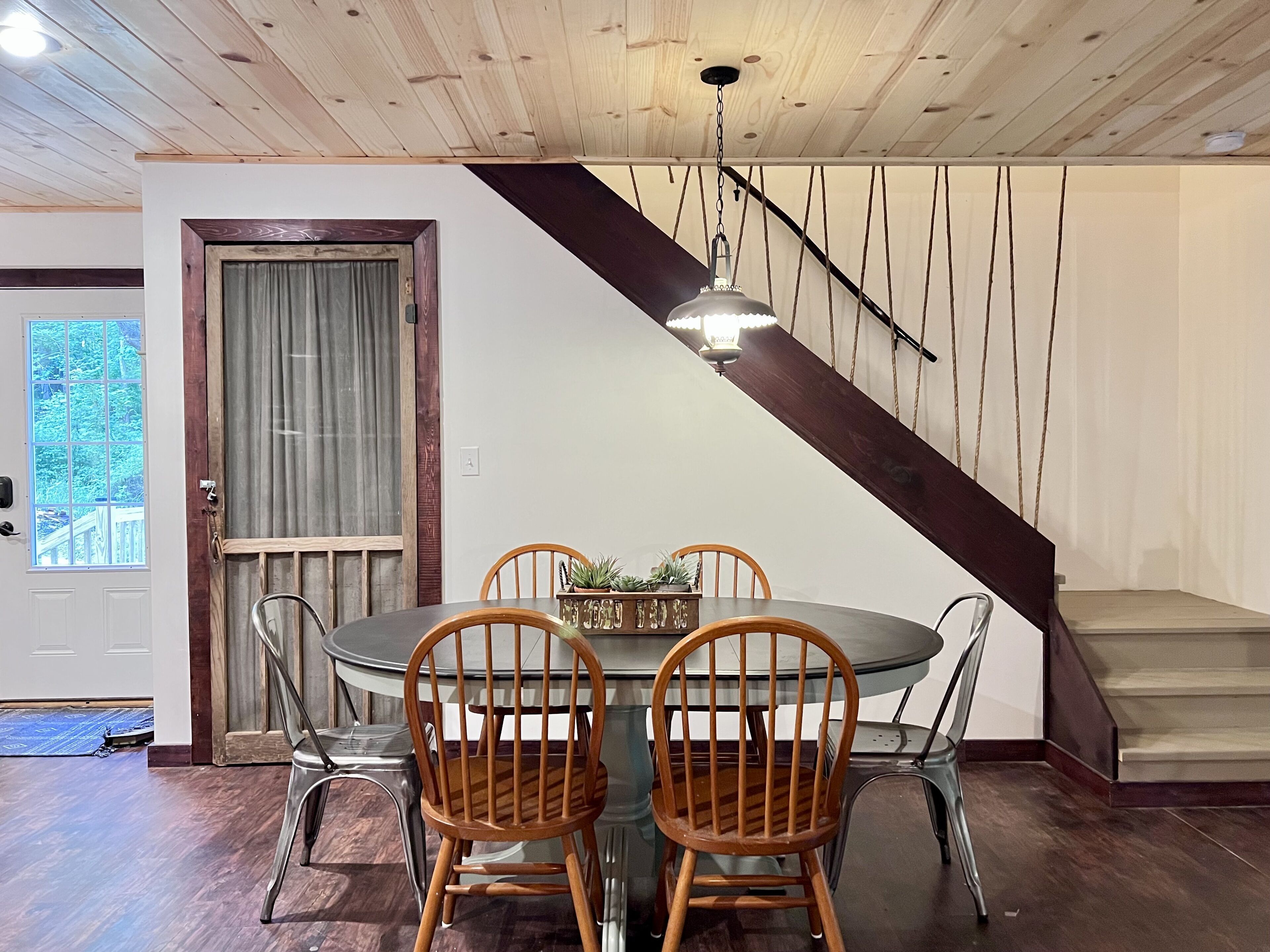 A dining area with a round table, four wooden chairs, a chandelier, and a staircase with a dark wooden handrail.
